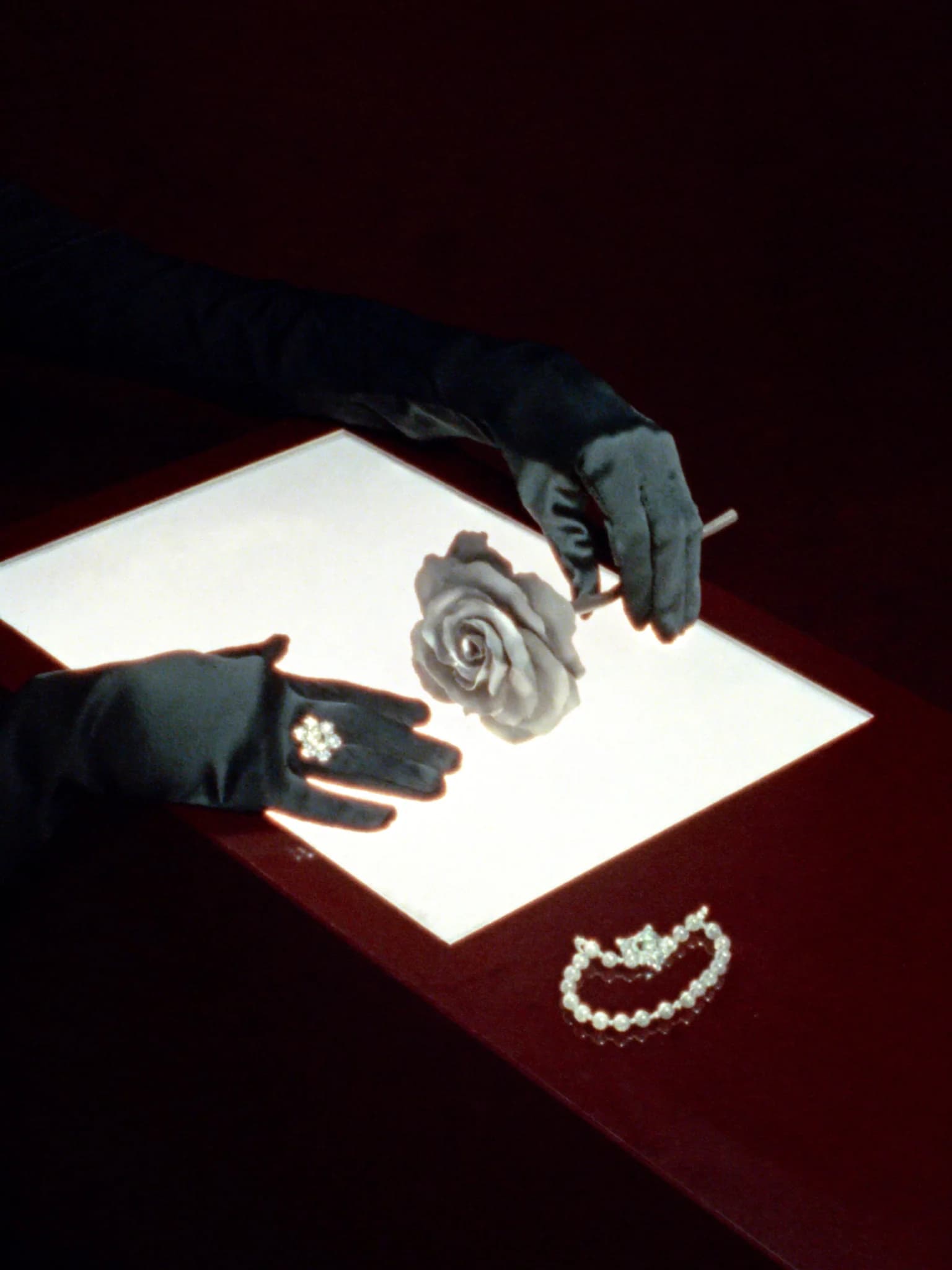 Black-gloved hands arranging a silver flower and jewelry over an illuminated oxblood table